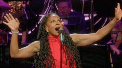 A Black woman with long wavy Black hair wearing a red sleeveless dress has her arms raised as she sings into a microphone