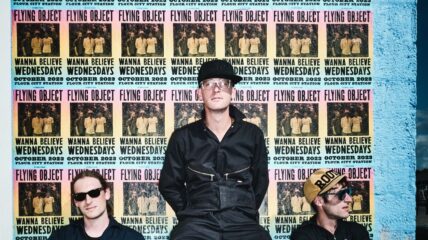 Three men in Flying Object Band Sit by poster wall
