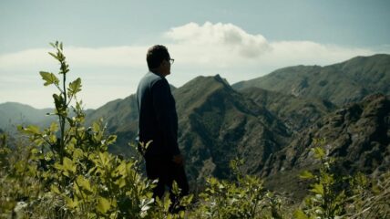 A white male stands in a field with mountains in the background.