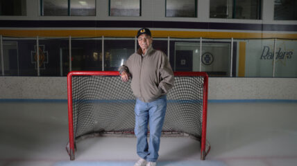 Henry Boucha wearing a tan coat, jeans and a baseball cop standing in front of a goal on an ice rink.
