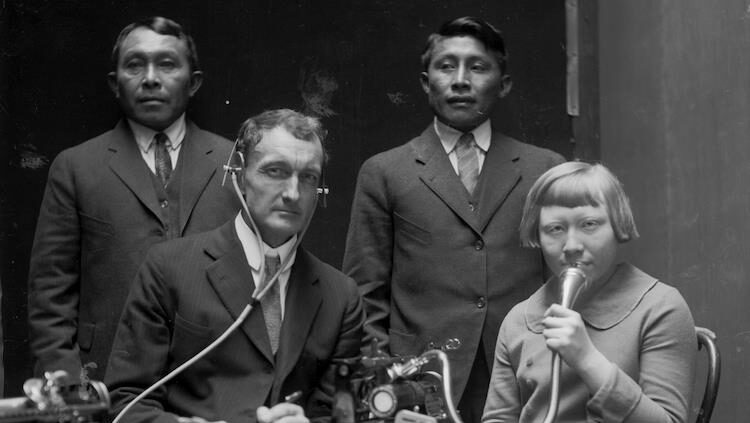 Four people in a room - three males and one female. Two men stand behind a man seated with stethoscope sits next to a women holding a microphone.