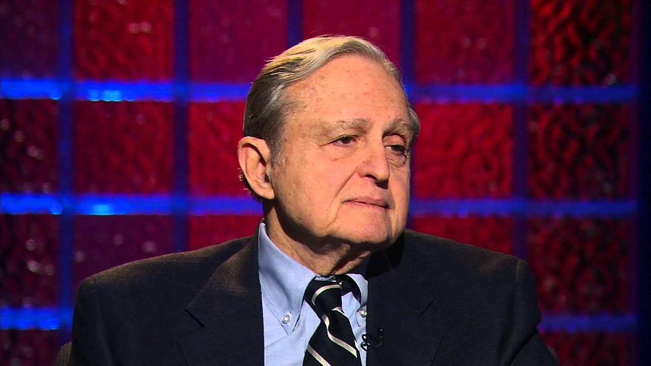 Attorney Justin Vigdor, a white older gentleman in a dark suite with a blue shirt and Black and silver striped tie sits on a TV set.