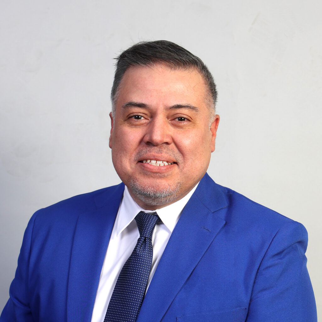 A Latino male with black short hair wearing a blue blazer, white shirt and dark blue tie.