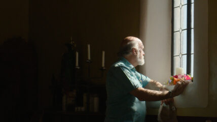 An older white man with gray hair stands in front of a window/