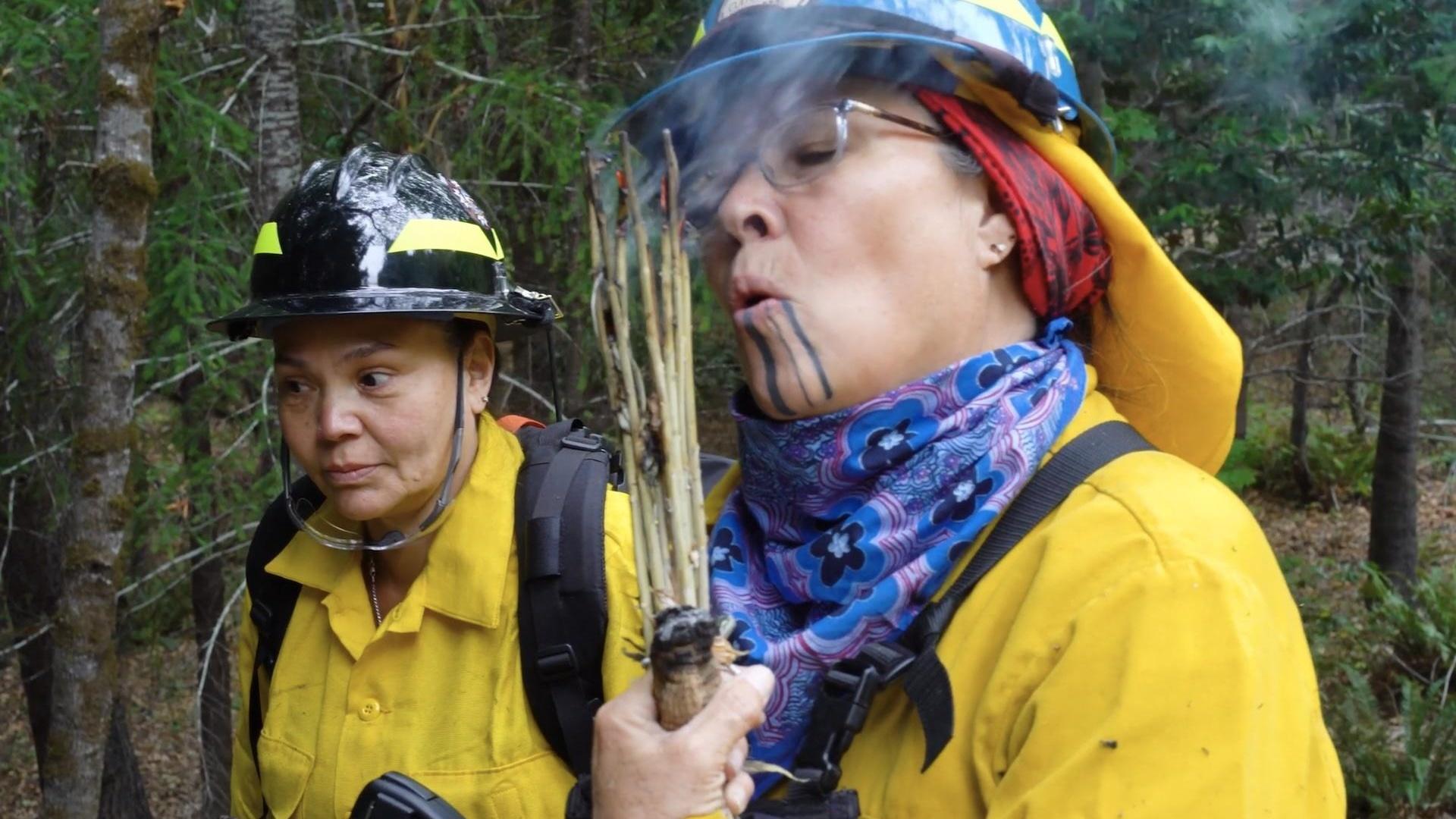 Two women firelighters stand next to each other. One women is holding a bunch of sticks together