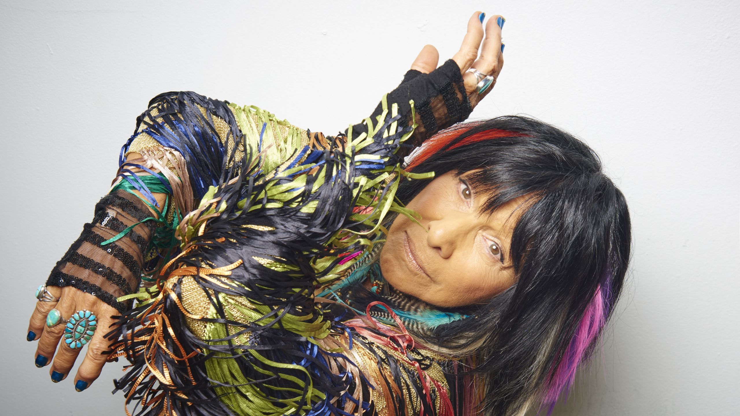 Buffy Sainte-Marie - shoulder length Black hair with colorful features weaved in. She is wearing a colorful jacket