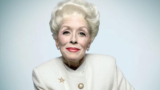 Actor Holland Taylor portraying Ann Richards. She has gray hair pulled back and up, wearing red lipstick and a white suit.