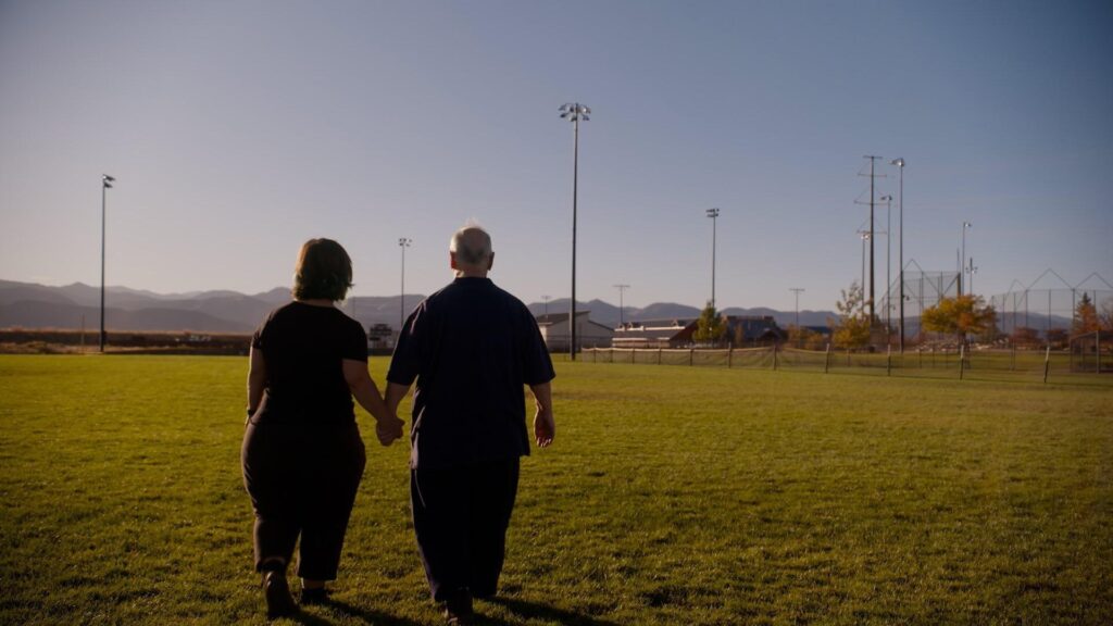 The filmmaker and her dad holding hands walking across a sports field away from the camera.