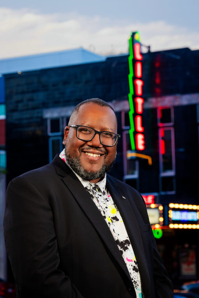 WXXI President Chris Hastings wearing a white shirt with splashes of paint and a black blazer. He is standing in front of The Little Theatre