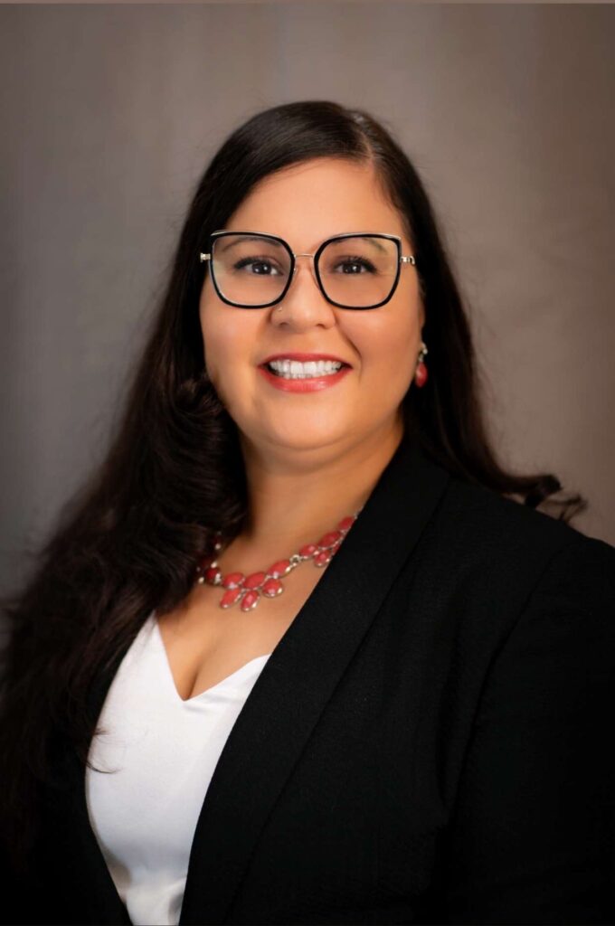 A Latinx woman with black shoulder length hair wearing black rimmed glasses, a black blazer and white blouse with a red beaded necklace.