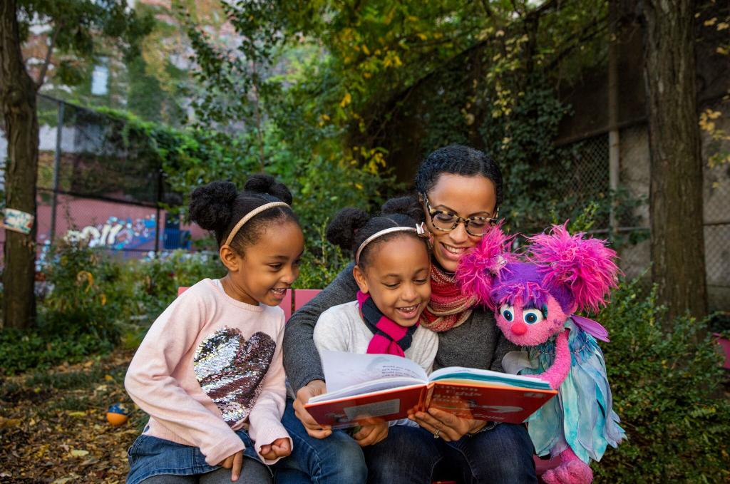 Mom and two children reading a book in a park setting with Abbey Cadabbey muppet from Sesame Street