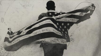 Black man facing away wrapped in a U.S. flag with some victims names written on the inside of it that have died due to racism and display backwards on the flag.