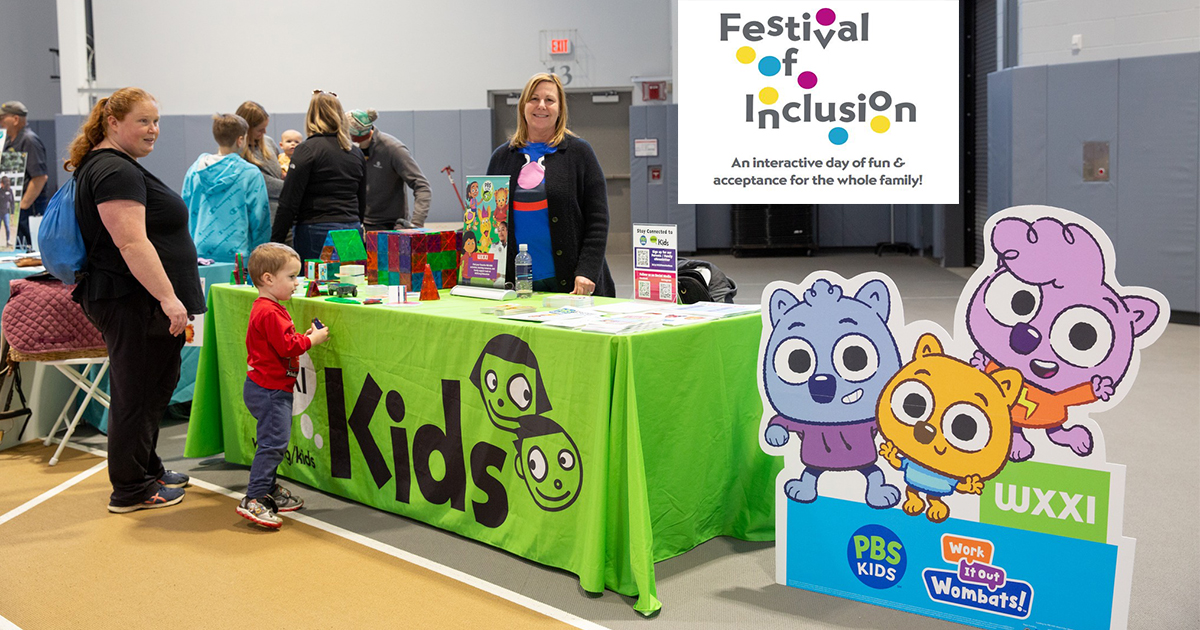 A mom and song stand in front of a WXXI Kids tableclothed table playing with building blocks.