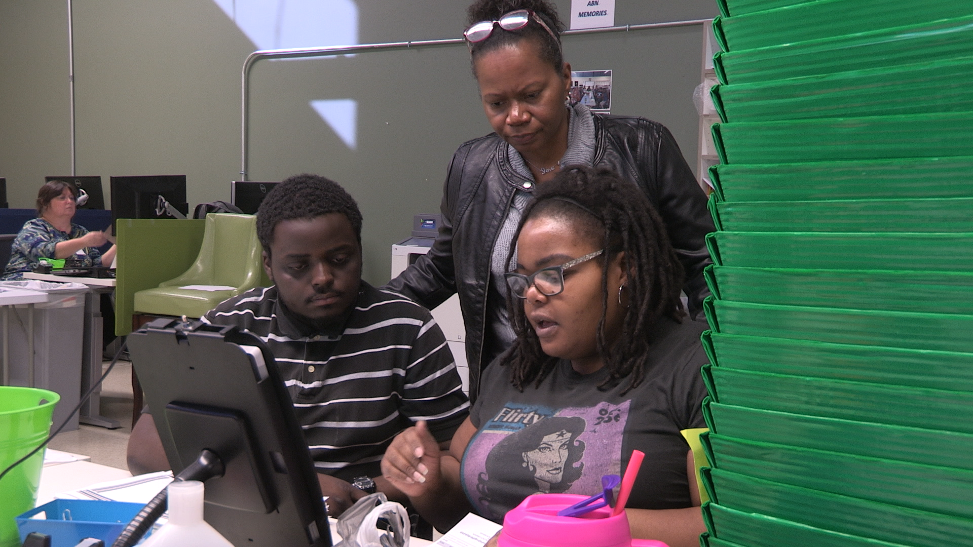 A young Black male in a stripped shirt sits next to his classmate, a young Black female who are working on the computer.