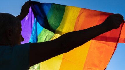 Women holding up a Pride Flag against a Blue Sky