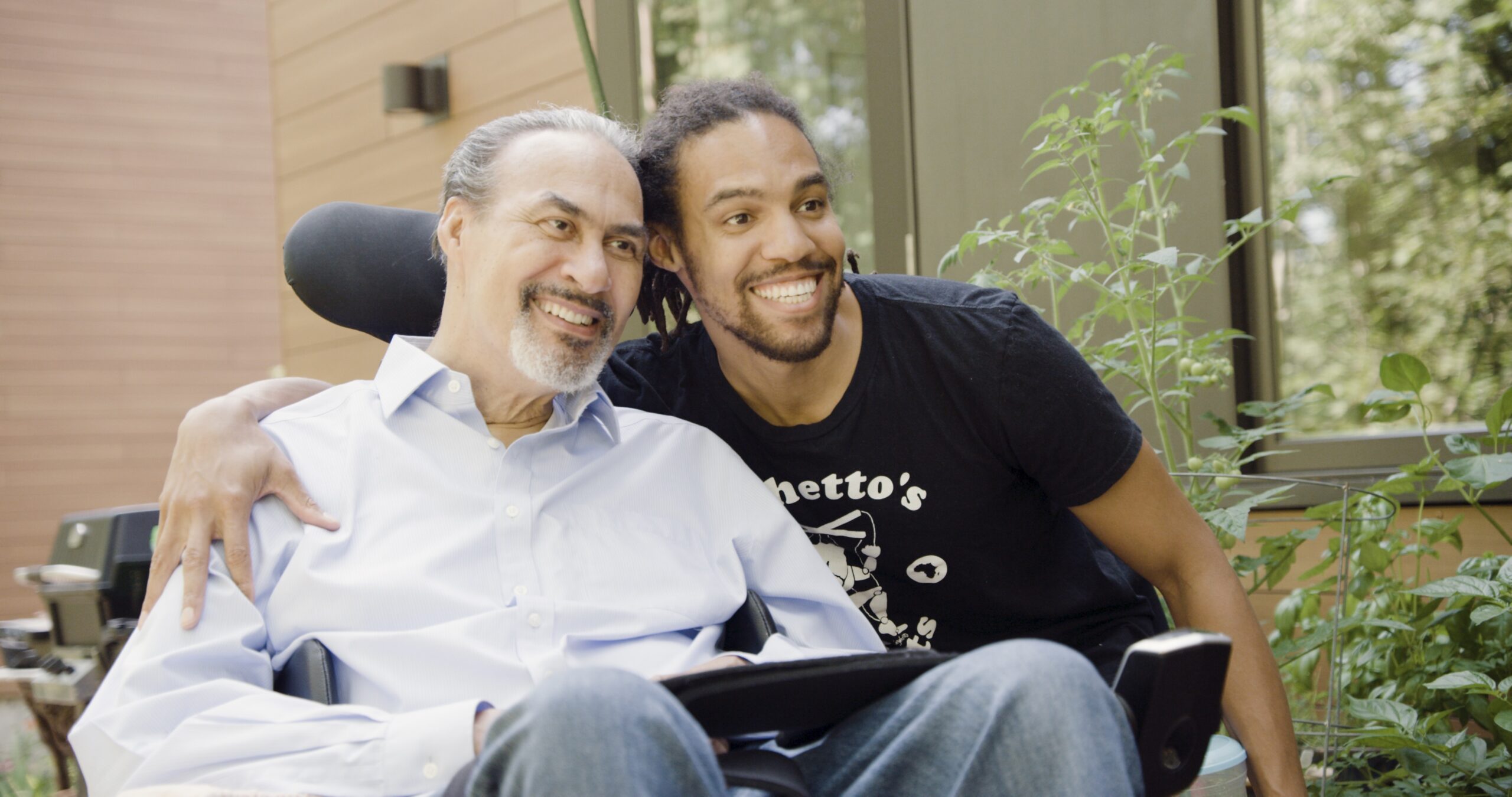 Two men smile. One with salt and pepper hair and beard and mustache wearing a white button down shirt. Second is a younger Black make with Black hair and beard and mustache.