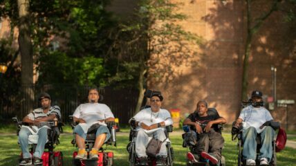 Five men in wheel chairs outside in front of a brick building.