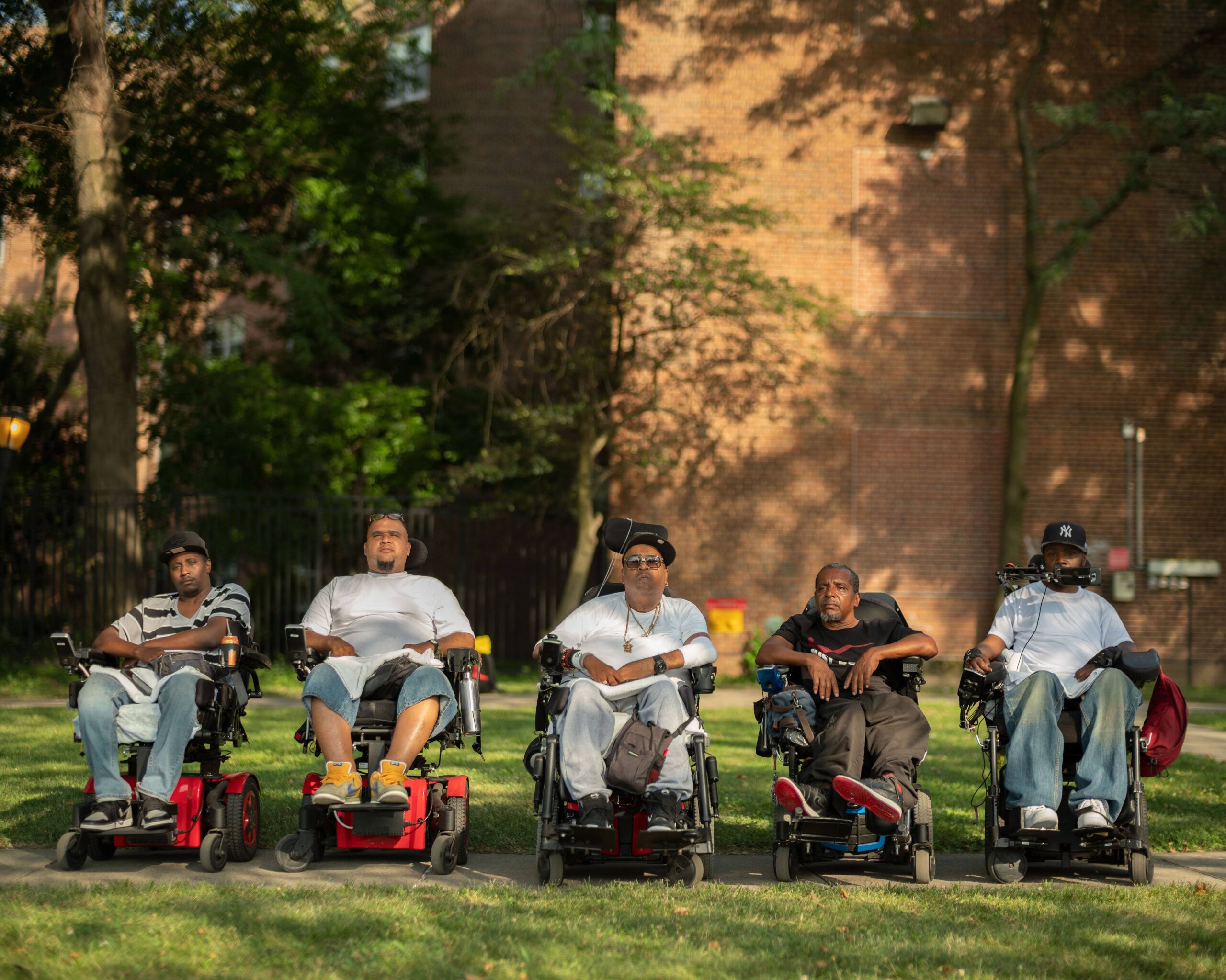 Five men in wheel chairs outside in front of a brick building.
