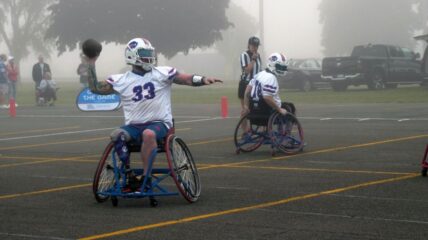 Two football players in wheel chairs - the one on the right drops back so that his teammate on the left can make a pass.
