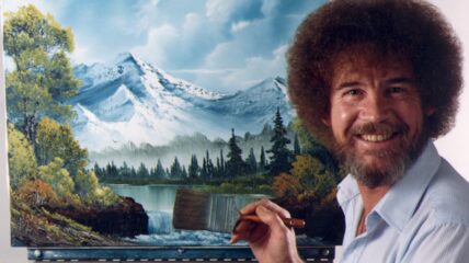 A white man with a brown afro, beard and mustache wearing a little blue button down shirt holding a pint brush in front of a canvass pairing a mountain and lake scene.