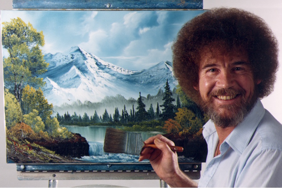 A white man with a brown afro, beard and mustache wearing a little blue button down shirt holding a pint brush in front of a canvass pairing a mountain and lake scene.
