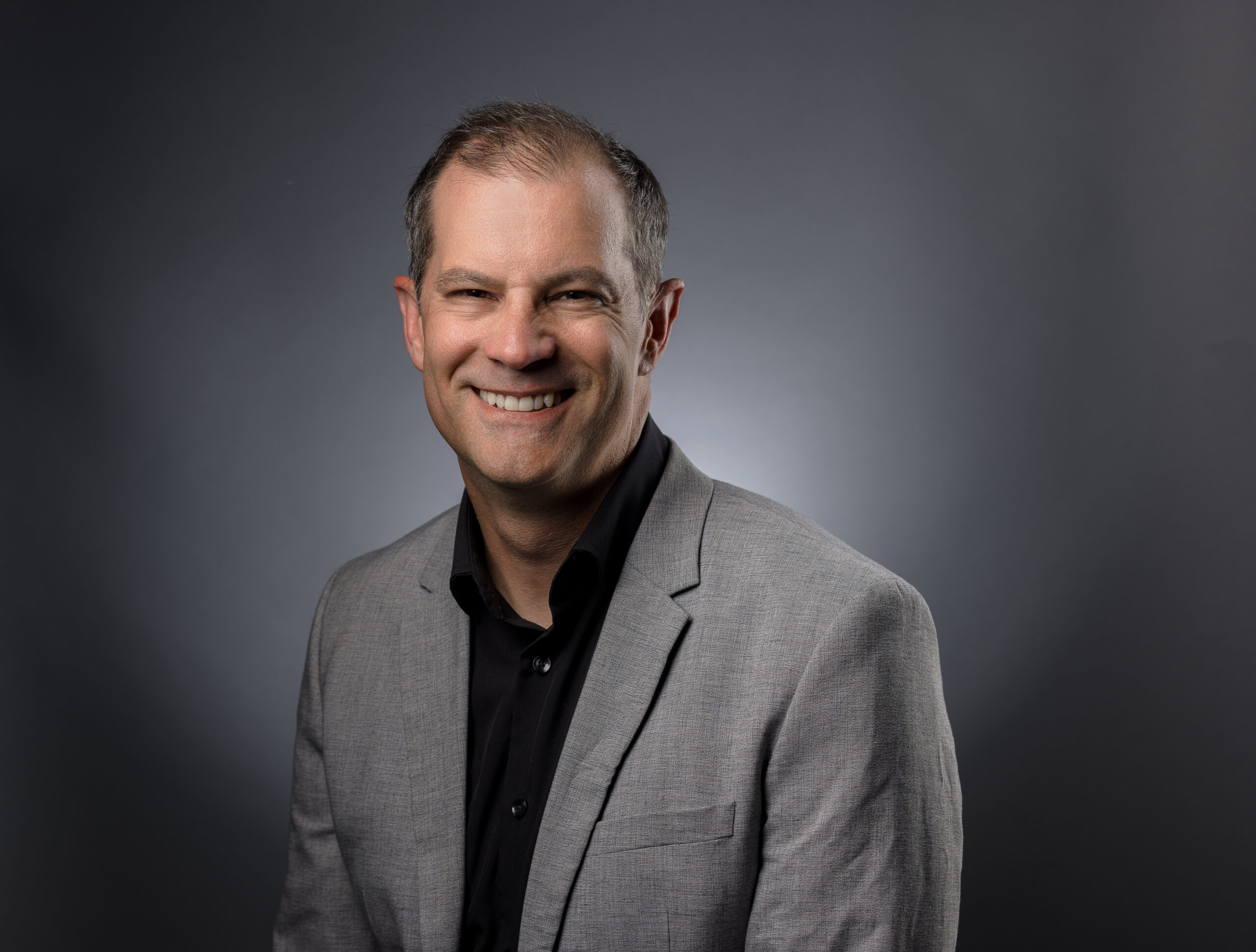 A white male with dark brown short hair wearing a dark blue button down and gray suit