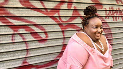 A Black woman with Black hair pulled up into a bun wearing a pink blouse stands in front of a silver garage door with graffiti.