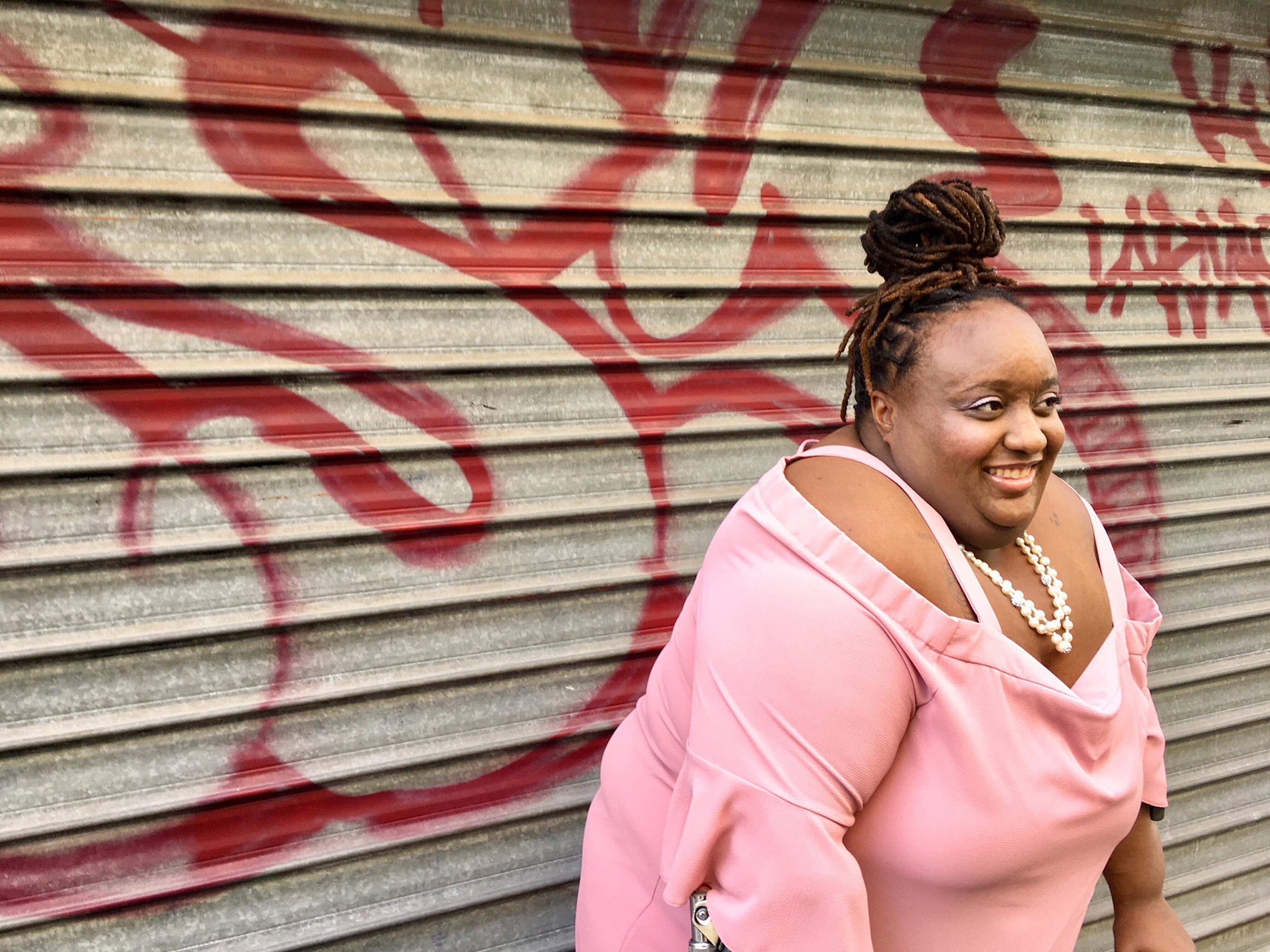 A Black woman with Black hair pulled up into a bun wearing a pink blouse stands in front of a silver garage door with graffiti.