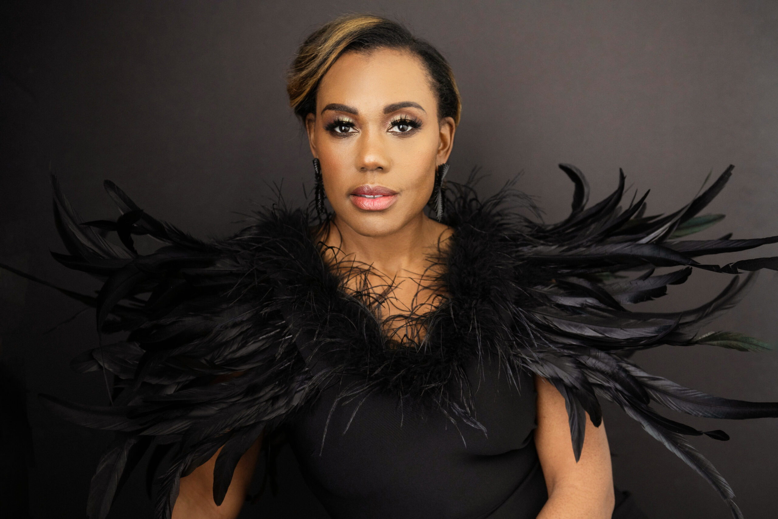 A Black woman with Black hair pulled back wearing a Black dress with a Black feathered boa.