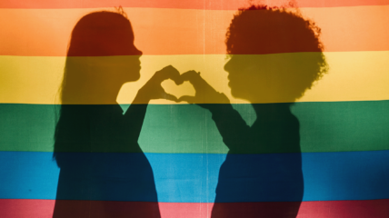 Silhouette of two people behind a rainbow flag forming a heart with their hands together each making half