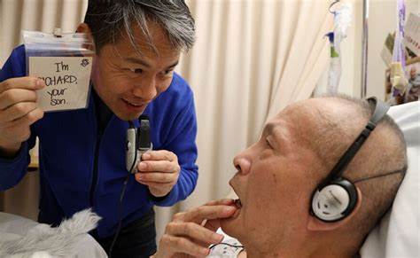 An Asian man with salt and pepper hair wearing a blue shirt holds an index card up to his elderly father who is in a hospital bed wearing headphones.