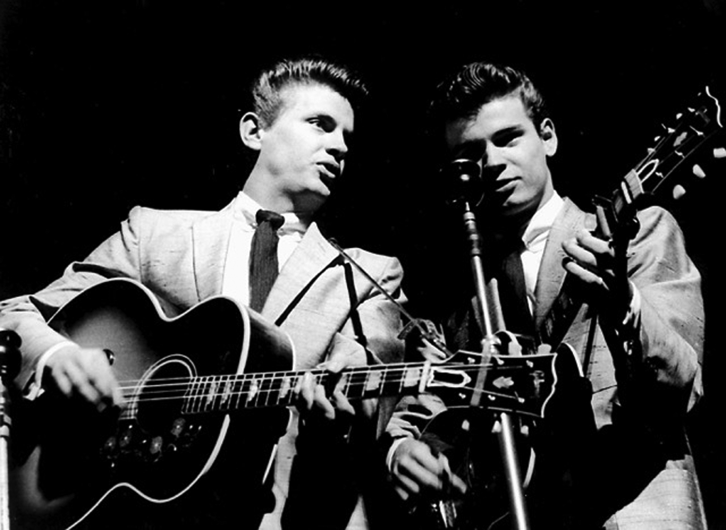 Black and white photo of the Everly Brothers. Both in matching suits. button down shirts and dark ties. Both playing guitar into a mic.