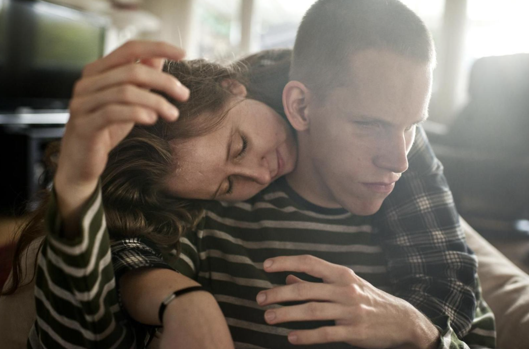 A white male wearing a striped t-shirt being hugged by his sister.