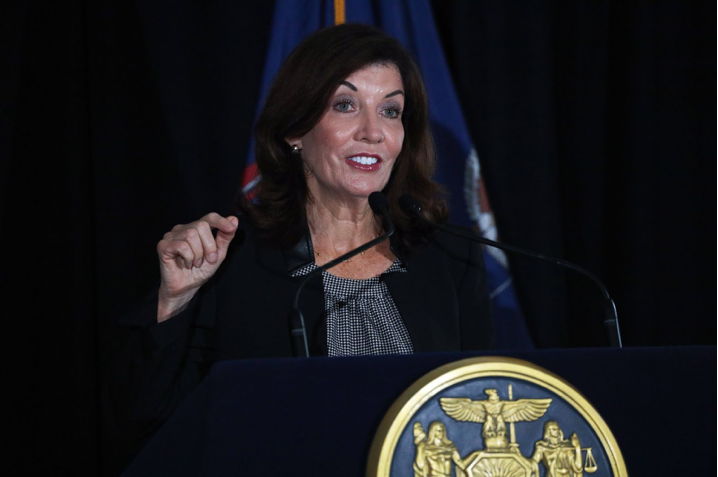 A white woman with long dark brown hair in a navy blue suit and gray blouse stands a a podium giving a speech.