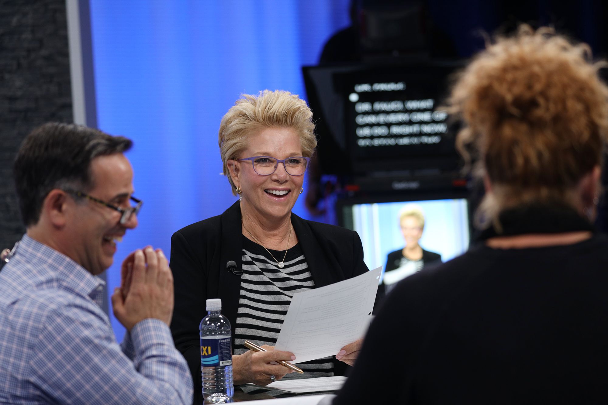 A white woman with short blonde hair gray rimmed glasses, a Black and white shirt with Black blazer smiles at at the woman across from her. A man sits in the middle of the two laughing. He has short Black hair and is wearing glasses.