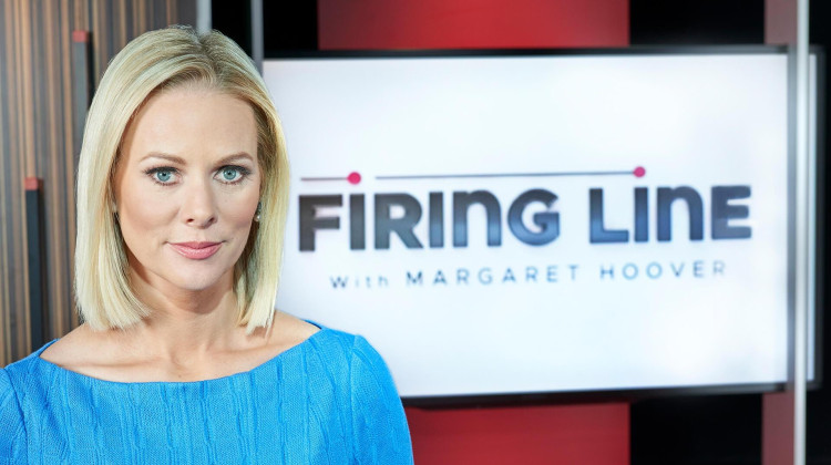 A white woman with long blonde hair and blue eyes, wearing a light blue blouse stands in front of a sign that reads: Firing Line with Margaret Hoover.