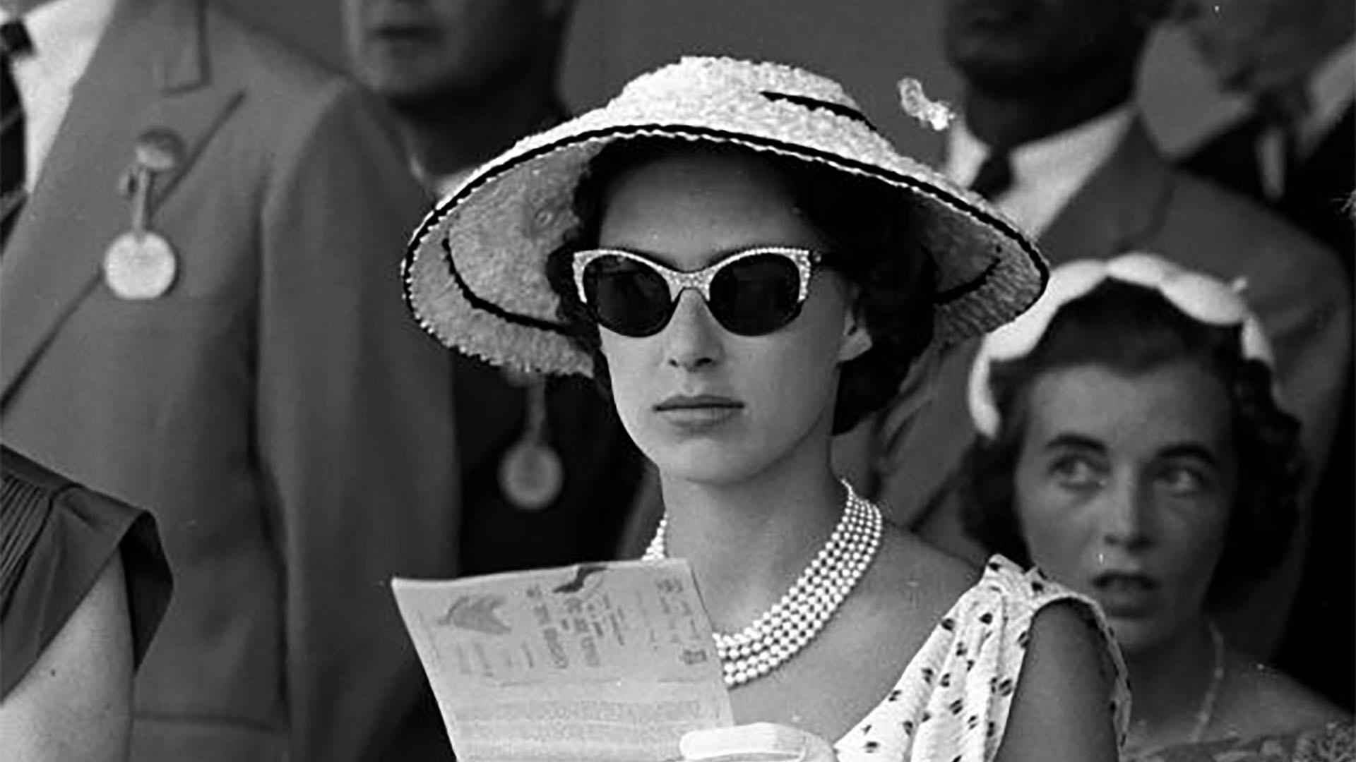 A Black and white photo of a young Princess Margaret wearing a white hat and sun glasses with pearls and a polk-a-dot dress.