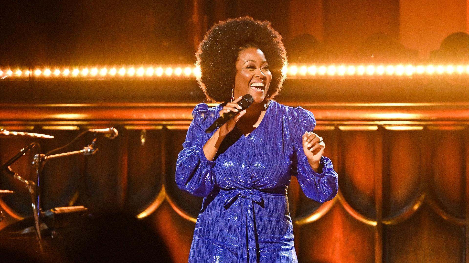 A Black woman with Black curly short hair in a blue dress sings on stage.