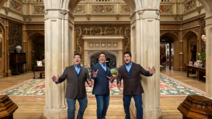 Three white men in dark blazers and jeans stand in front of a great room in a castle with their arms outstretched