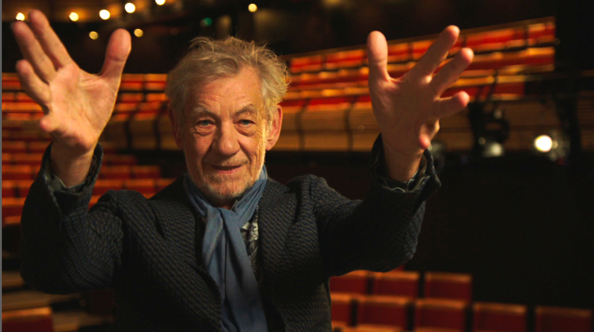 A white, older man with gray hair holds his hands up in the air. He's wearing a gray cardigan and a blue scraf
