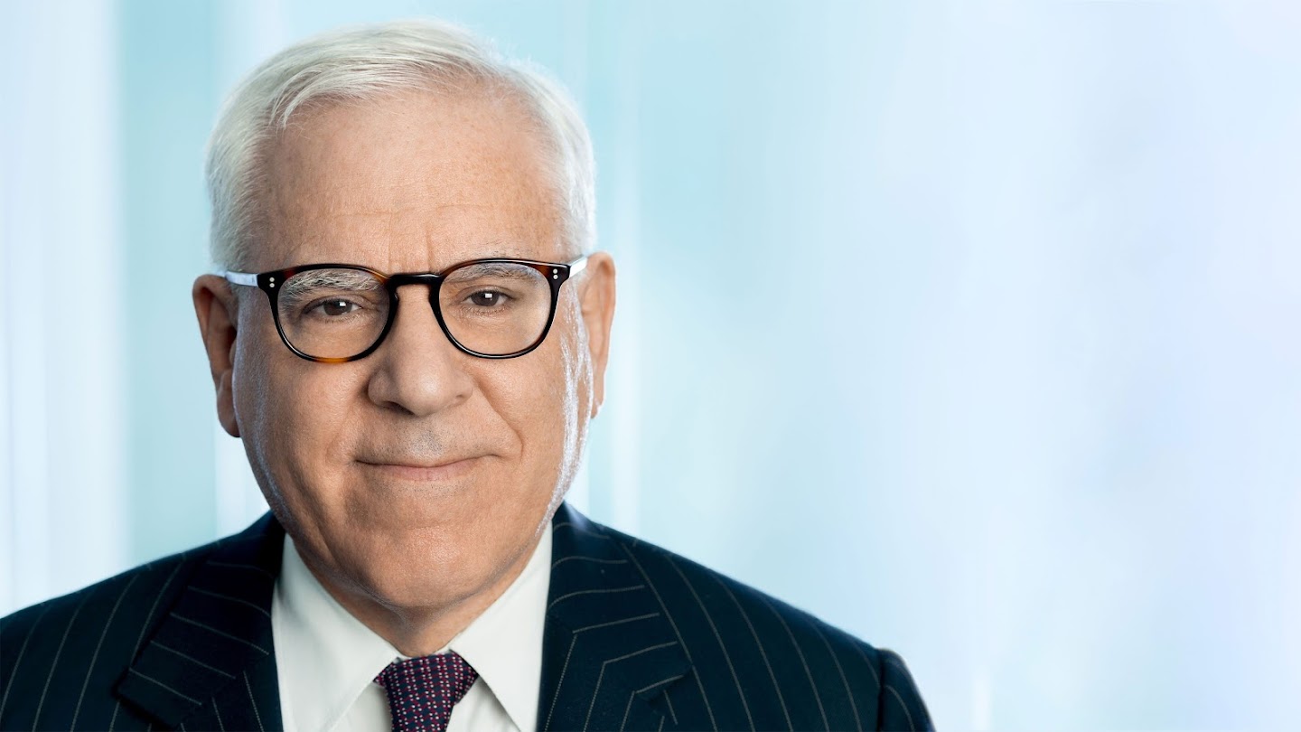 A photo of David Rubenstein - a white male with short gray hair wearing black rimmed glasses and a suit and tie.