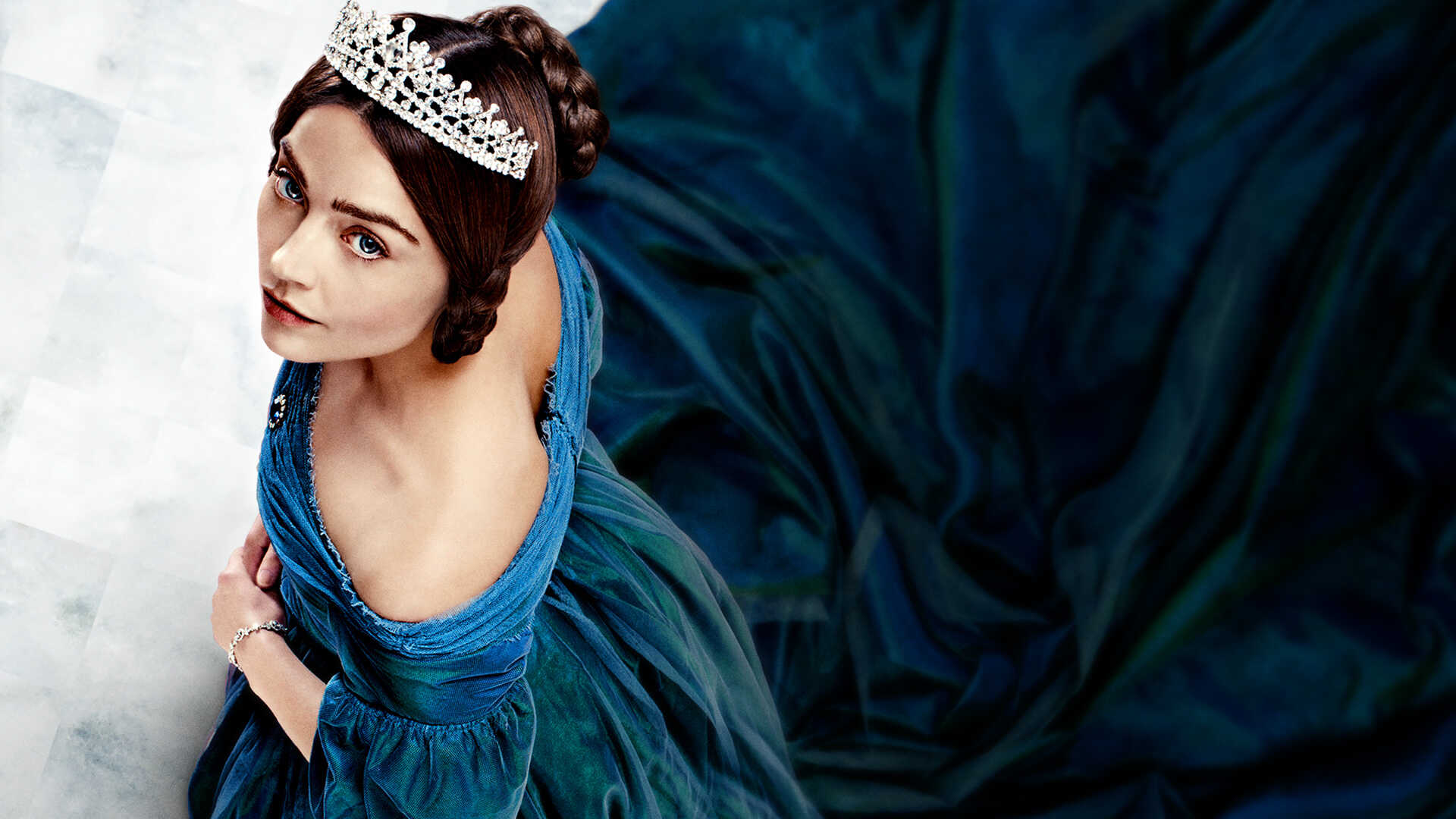 A white woman with brown hair pulled back wearing a diamond studded crown and a blue dress looks up at the camera.