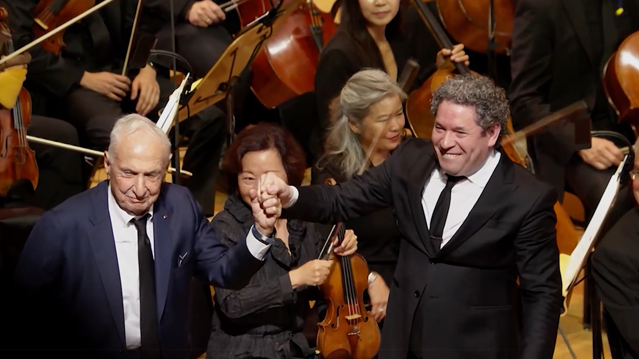 Three men on a stage with a full orchestra behind them. Two of the men are holding hands and smiling.