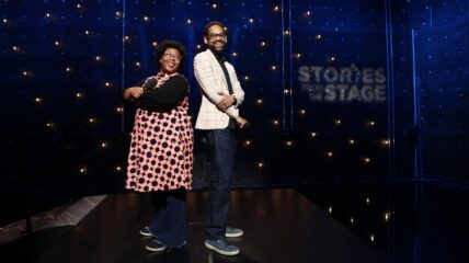 A Black woman and a Black man stand back to back holding mics on a stage