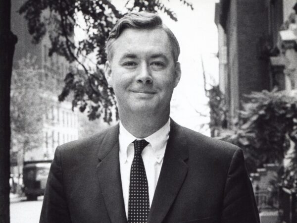 A Black and white picture of a white man with dark short hair, wearing a dark suite and tie, and a white button down shirt.