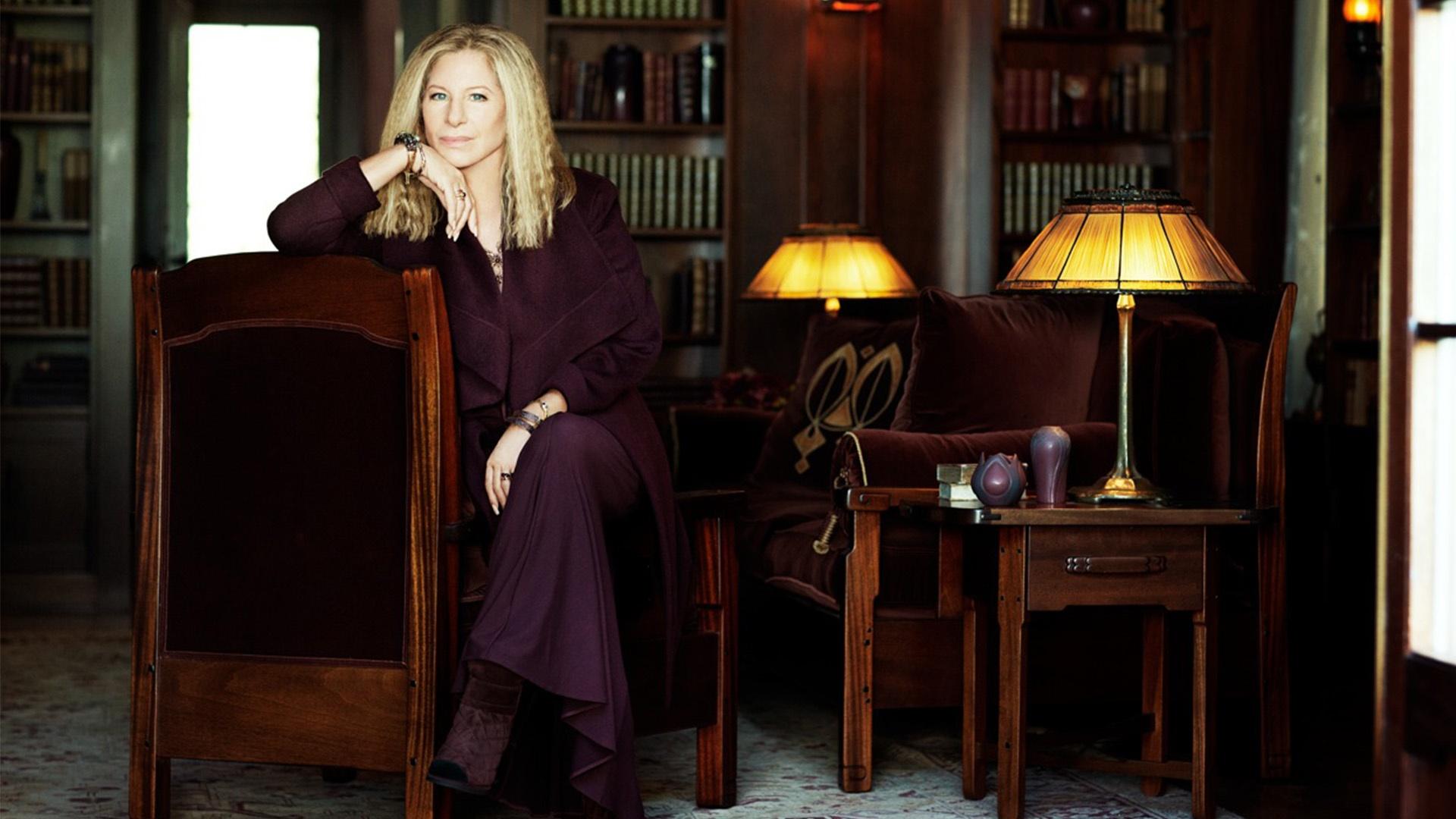 A white woman with long blonde hair wearing a marron colored dress sit in a chair with one arm folded over the top of the chair as her hand holds her chin