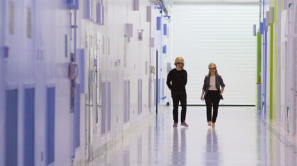 A white man and white woman wearing yellow hard hats walk down a lab hallway