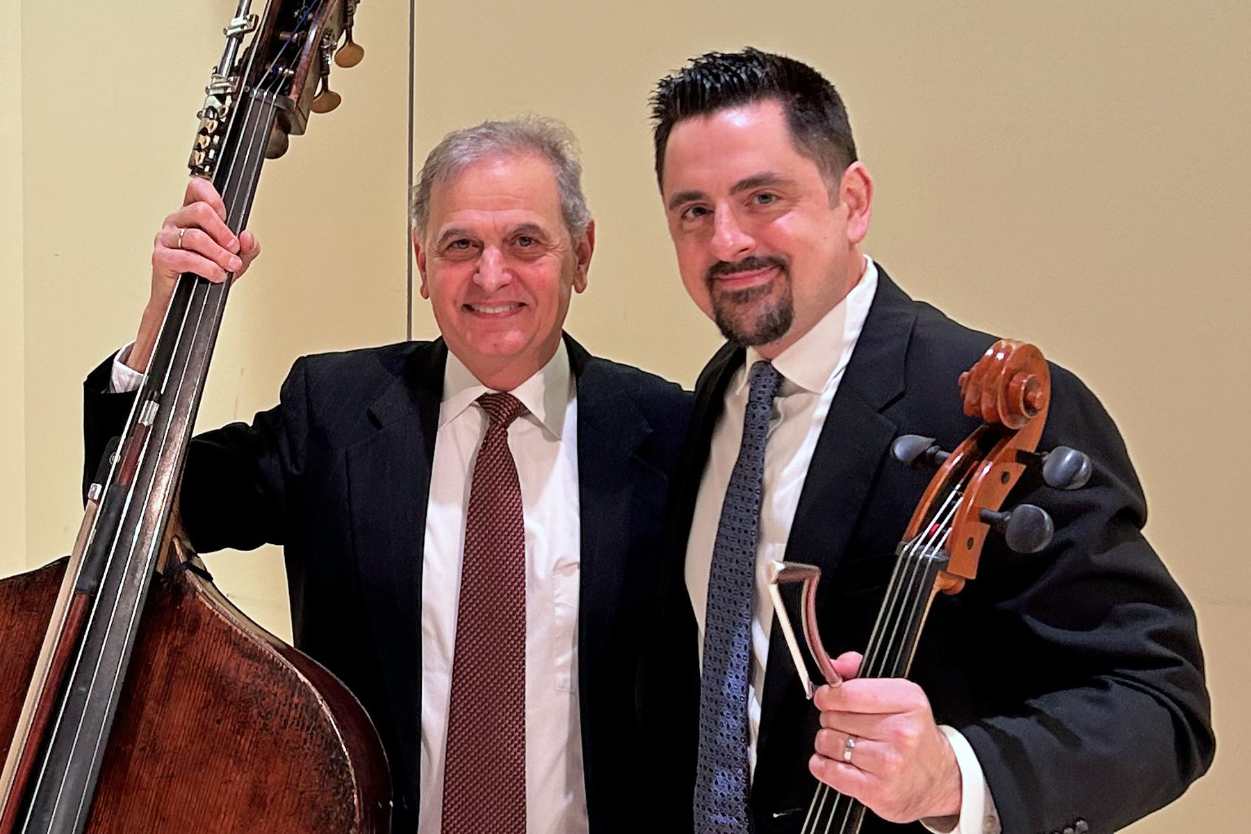 Two men stand together. The one on the left is a white man with gray hair and a white made in dark brown hair with a mustache and goatee. Both wearing dark suits, white collared shirt and ties.