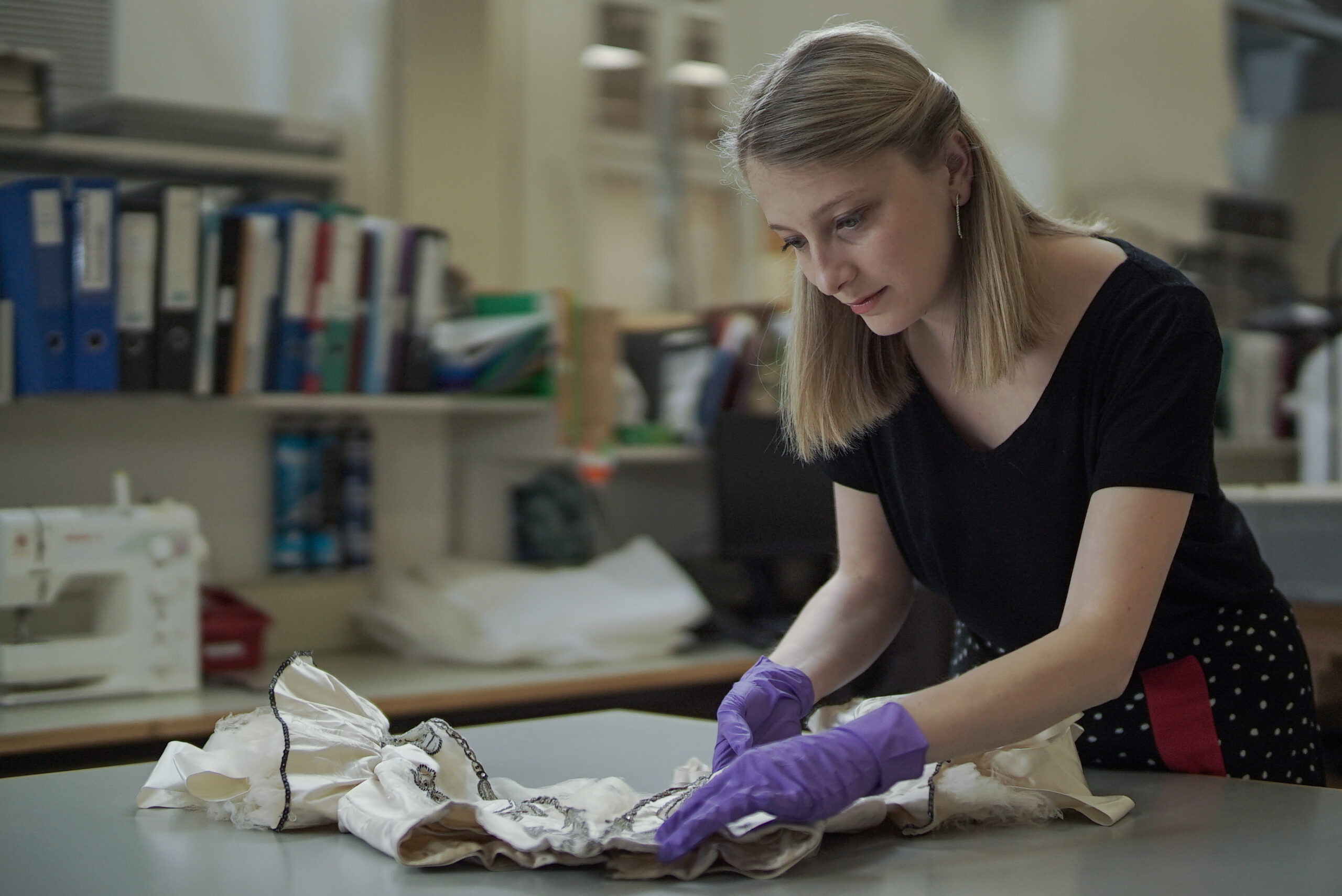 A white woman with long blonde hair wearing a Black t-shirt and purple plastic gloves holding some white fabric