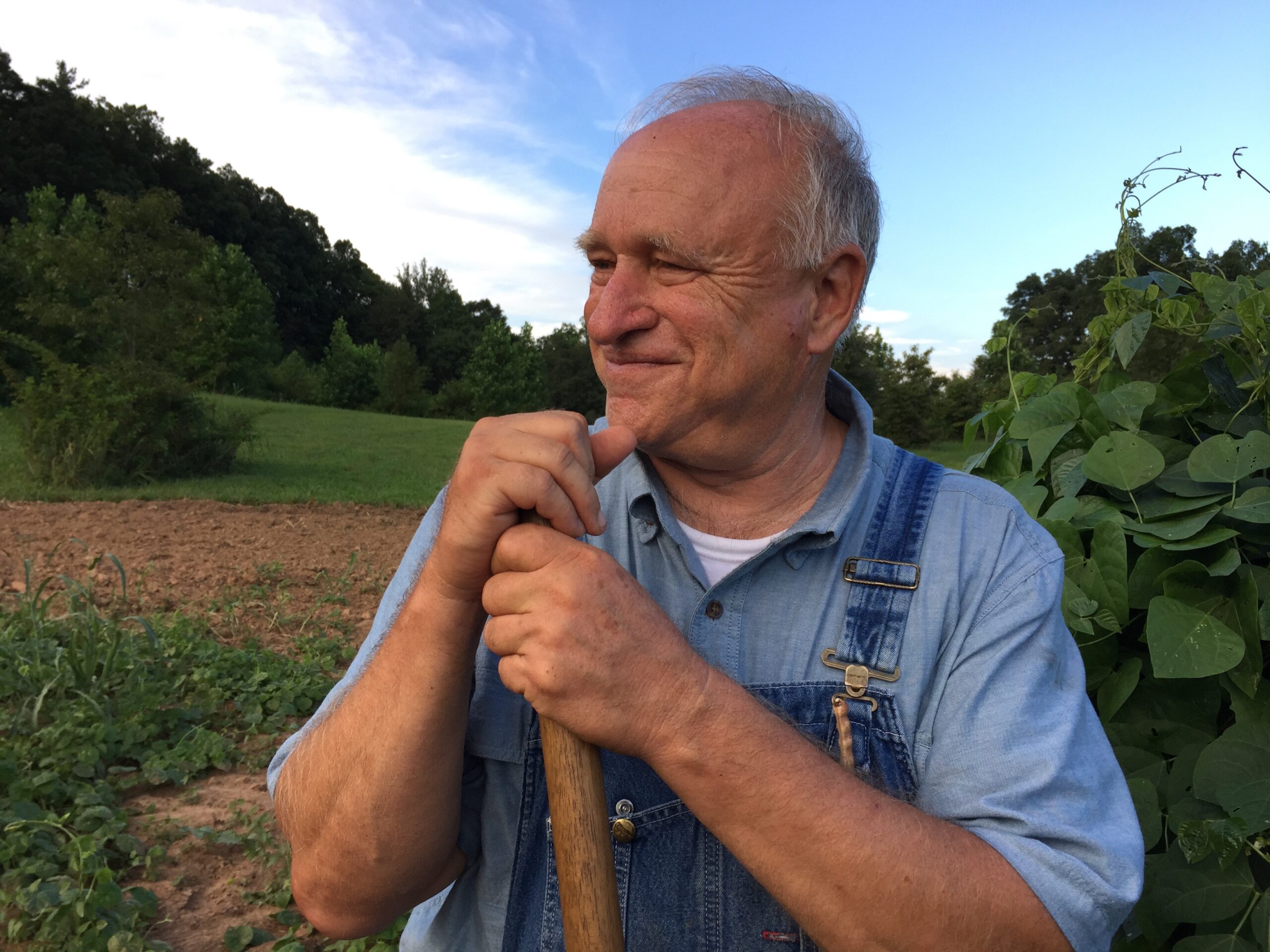 An older whit mad with short gray hair wearing a blue collared shirt + overalls has his hand resting on the handle of a rake.